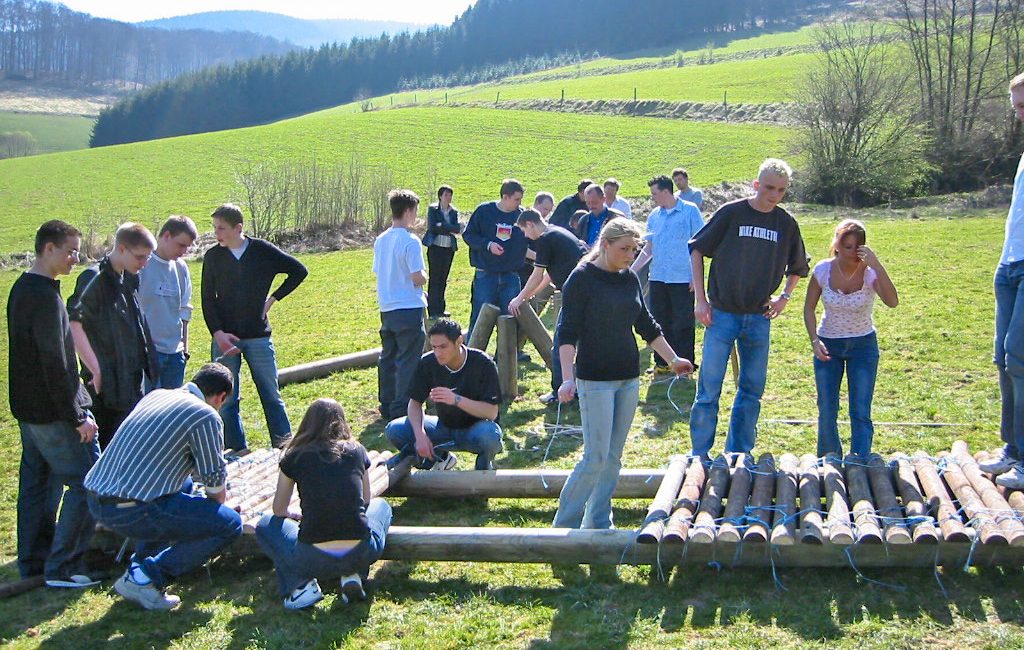 Gruppe bei Outdoor-Teamübung im Sauerland auf einer Wiese, konstruiert ein Floß aus Holz