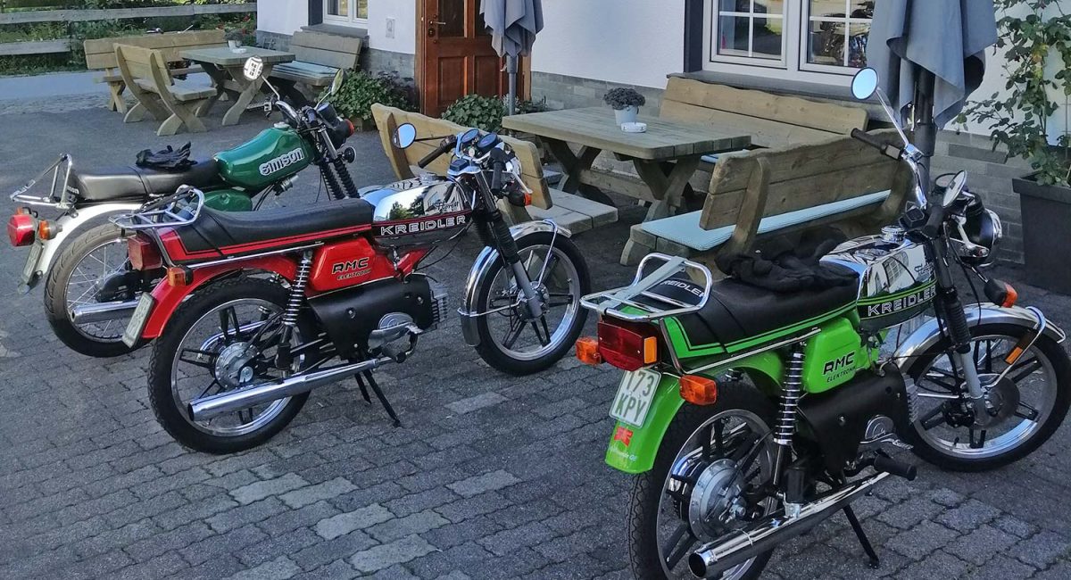 Mehrere Mopeds stehen nebeneinander auf einem Hof im Grünen